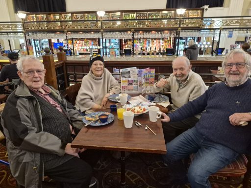 Four elderly friends seated at a table with breakfast items in a café setting.